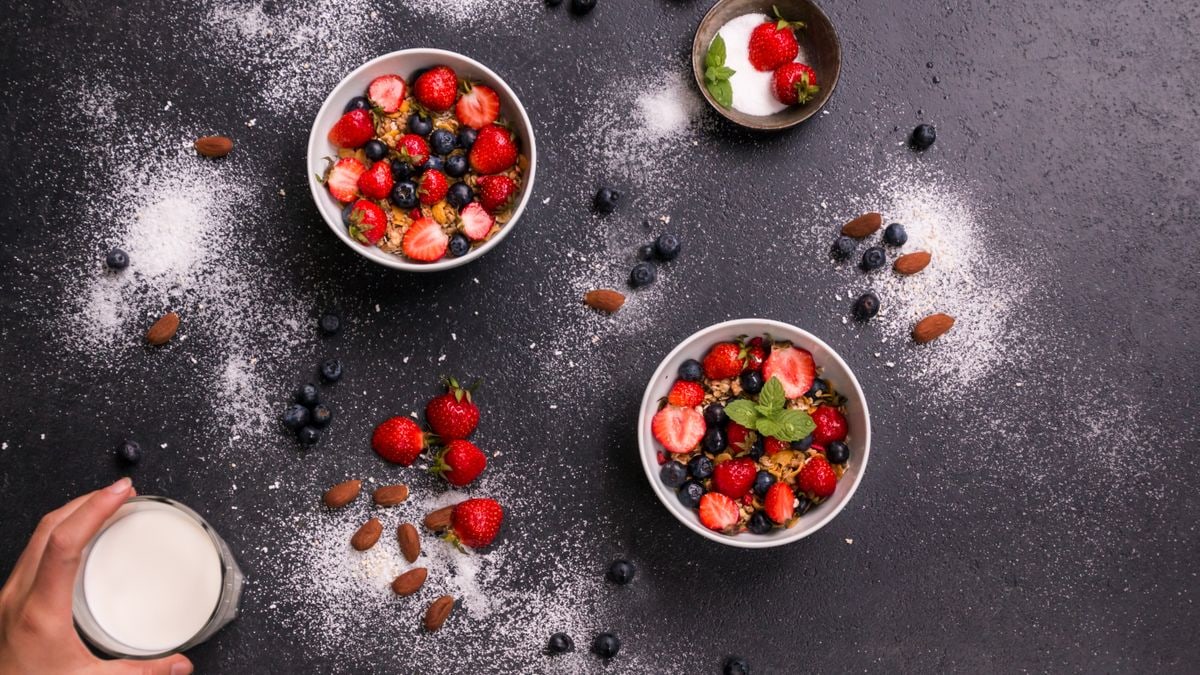 Tasty berry breakfast bowls with almonds and glass of milk on dark surface.